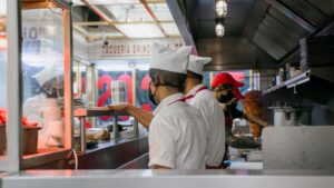 Two fast food kitchen workers preparing fried chicken at a stainless steel bench inside a small quick-service restaurant kitchen
