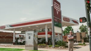 A woman holding fuel receipts stands next to a van at an Australian petrol station, looking concerned as she reviews the costs