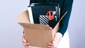 A woman in a bank office placing personal belongings into a cardboard box on her desk while a colleague looks on from a nearby workstation